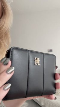 Portofel negru din piele cu accesorii aurii, având un design cusut și un logo proeminent pe partea din față. Închidere cu fermoar pe margine.