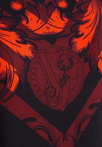 T-shirt Versace avec un design audacieux rouge et noir présentant des motifs complexes et un logo central. La texture semble lisse.