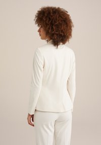 Blazer blanc ajusté avec une texture lisse, des manches longues et des détails de couture subtils, associé à un pantalon assorti.