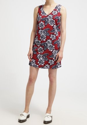 Ærmeløs rød minikjole med hvidt og sort blomstermønster og bladmotiv, båret med hvide loafers på en stående person.