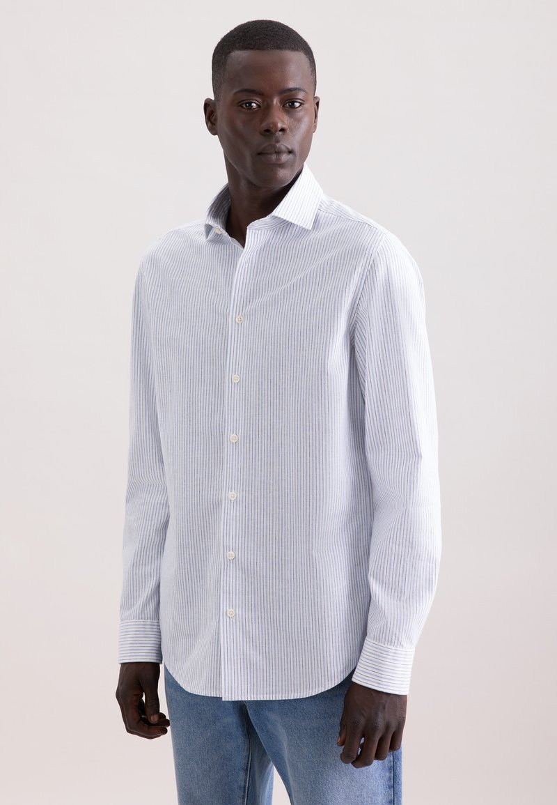 Homme portant une chemise blanche à manches longues avec de fines rayures verticales et un jean bleu, debout devant un fond clair uni.
