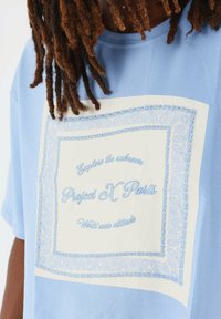 Camiseta de color azul claro con un diseño de paisley blanco y texto azul. Con mangas cortas, corte relajado y material de algodón suave.