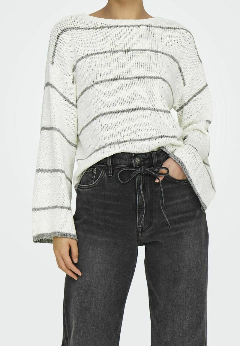 Personne portant un pull en maille blanc avec des rayures horizontales grises et un jean noir taille haute noué avec un nœud sur le devant.