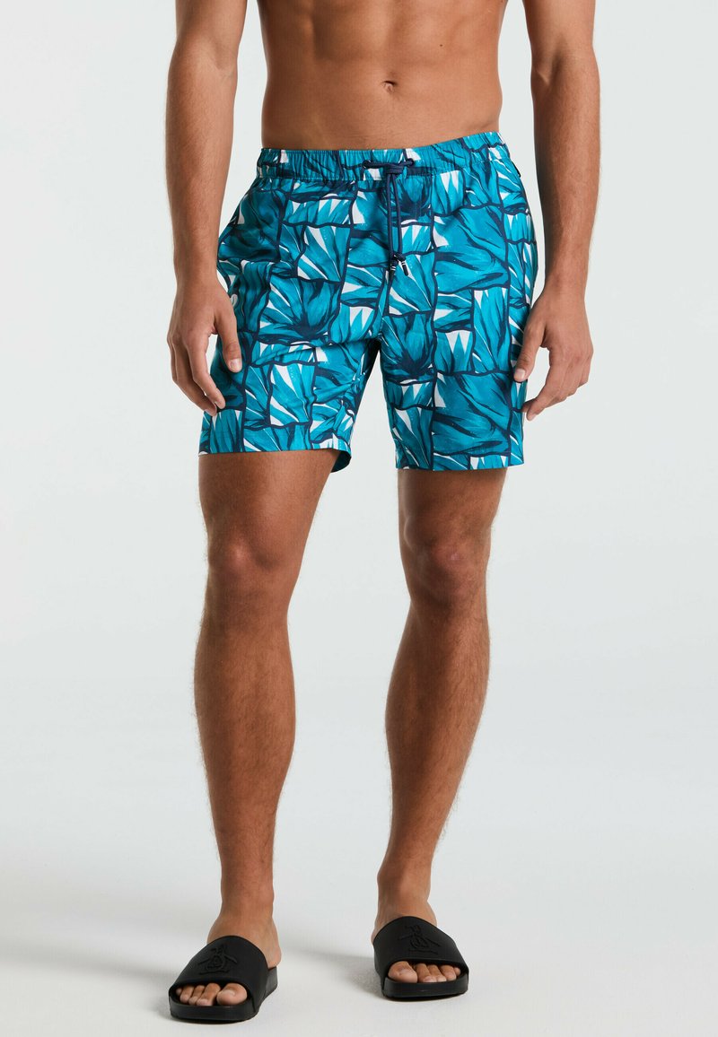 Un short de bain bleu présente un motif de feuilles tropicales dans des nuances variées, avec une taille élastique et un cordon de serrage. Associé à des sandales à glissière noires.