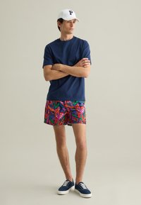 Camiseta azul marino, coloridos pantalones cortos de baño tropicales, gorra blanca con la letra "P" y zapatillas azul marino con cordones blancos.