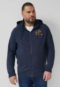 Marineblauwe hoodie met rits, voorzien van een capuchon met trekkoord, voorzakken, geribbelde manchetten en een grafisch logo op de borst. Soepele textuur.
