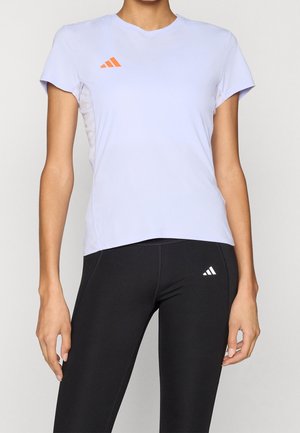 Femme portant un t-shirt de sport lavande à manches courtes avec un logo orange et des leggings noirs avec un logo blanc, debout devant un fond uni.