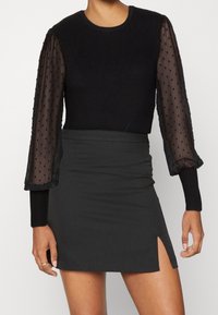 Top lungo a maniche ribbed nere con maniche puff trasparenti a pois, abbinato a una mini gonna nera con uno spacco laterale.