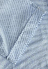 Gaiši zila kokvilnas auduma ar plānām baltām svītrām, gluda tekstūra, salocīts malas, un redzams rūpīgi izšūta šuve uz manžetes zonas.