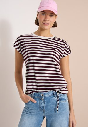 Giovane donna con cappellino rosa, maglietta a righe nere e bianche con fiocco laterale e jeans azzurri chiari, in piedi con una mano in tasca.