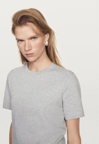 Szary t-shirt z krótkim rękawem, wykonany z miękkiego materiału, z okrągłym dekoltem. Model ma proste włosy i nosi duży srebrny kolczyk. Tło jest białe.