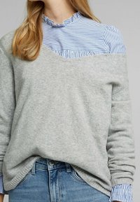 Femme portant un pull gris clair épaules dénudées sur un chemisier à volants rayé bleu et blanc, associé à un jean bleu, raccourci au niveau du cou et de la taille.