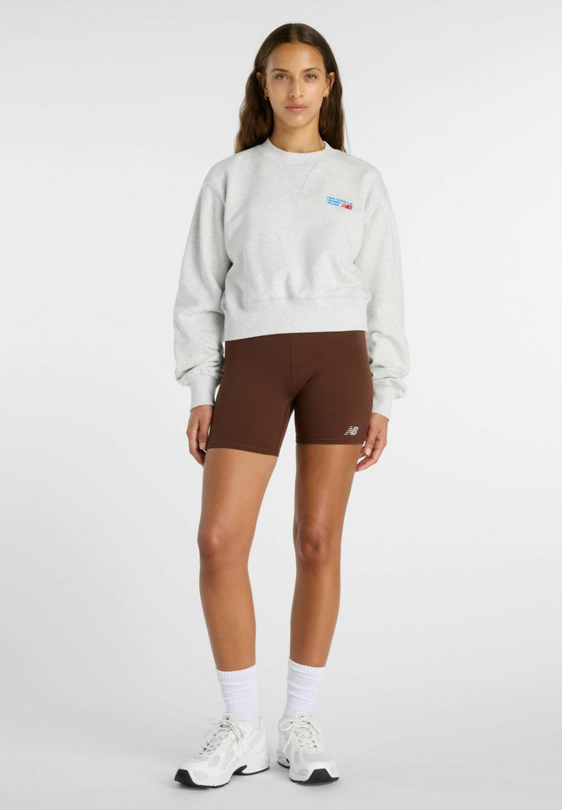 Jonge vrouw die een lichtgrijze trui, bruine nauwsluitende korte broek, witte sokken en witte sportschoenen draagt, staand tegen een effen achtergrond.