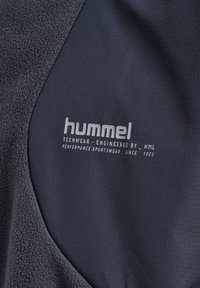 Mörkgrå techwear-tyg med en texturerad, fleeceaktig sektion, med "hummel"-logotyp och detaljer för prestationssportkläder i vitt sydda.
