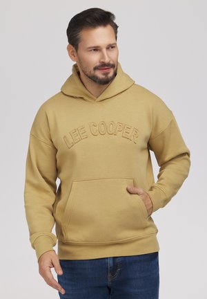 Mężczyzna z brodą w beżowej bluzie z kapturem Lee Cooper i niebieskich dżinsach, prawą ręką w kieszeni, lekko patrzący w lewo na jednolitym tle.