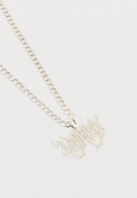 Ασημένιο κολιέ με αλυσίδα και μενταγιόν σε σχήμα φλόγας που διαθέτει αφηρημένο σχέδιο με κοφτερές, αιχμηρές εγκοπές σε απλό ανοιχτό φόντο.