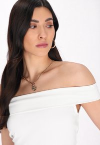 Hvid off-shoulder top i glat stof med en subtil glans og en sideslids. En halskæde med en cirkulær vedhæng hviler elegant på kravebenet.