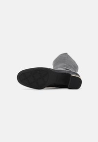 Bota de cuero negro hasta la rodilla con punta afilada, suela texturizada y un sutil acento dorado en el tacón. Parte superior suave y construcción robusta.