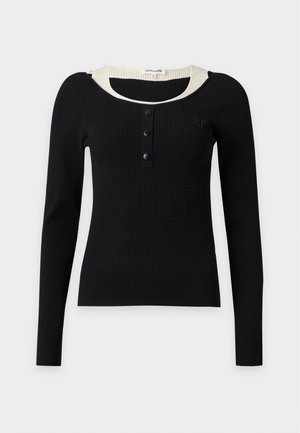 Ung person med kort hår iført en sort langærmet top, blå vide jeans og sorte spidse sko, stående med den ene hånd i lommen.