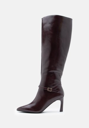 Botas castanhas de couro até ao joelho com bico pontiagudo, salto agulha e uma tira decorativa com fivela à volta do tornozelo. Textura lisa.