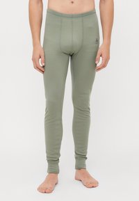 Uomo che indossa pantaloni interi aderenti verdi, in piedi a piedi nudi contro uno sfondo bianco semplice.