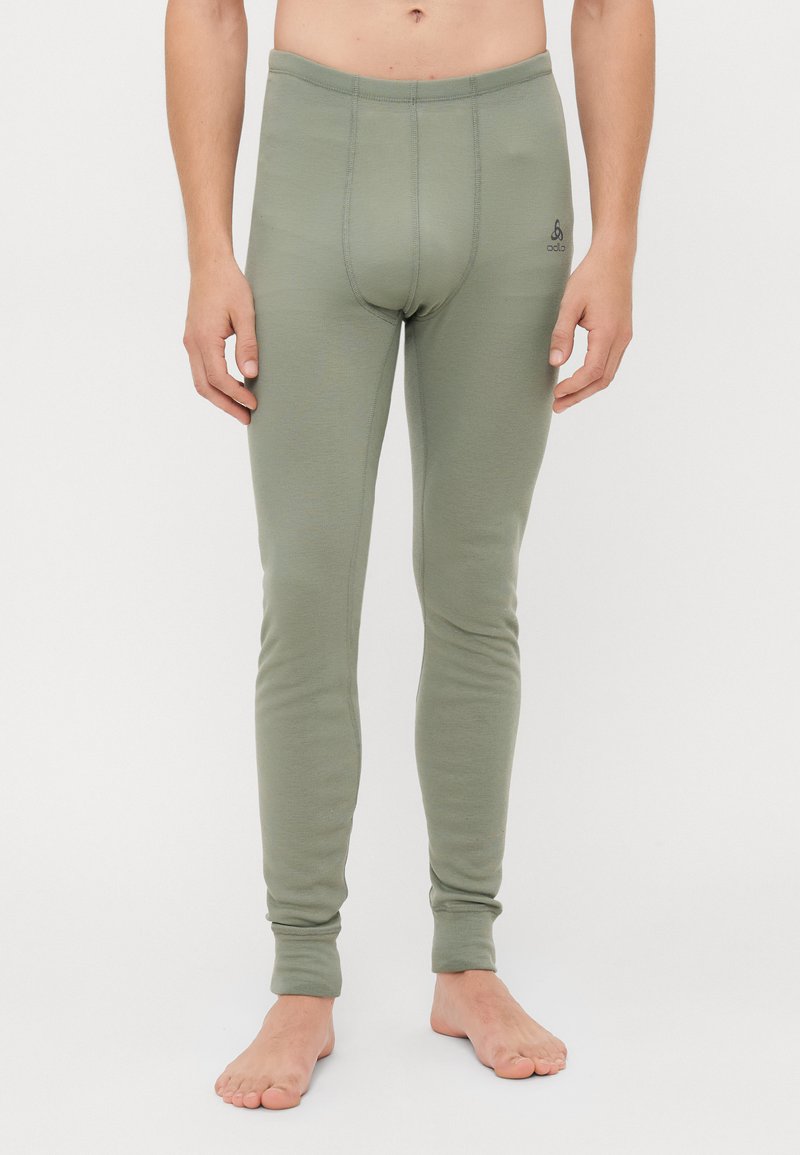 Uomo che indossa pantaloni interi aderenti verdi, in piedi a piedi nudi contro uno sfondo bianco semplice.