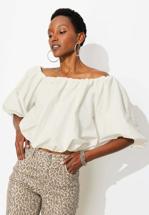 Personne aux cheveux courts portant de grandes boucles d'oreilles créoles, un top blanc à épaules dénudées et manches bouffantes, un pantalon imprimé léopard et un bracelet en argent, posant avec les mains dans les poches.