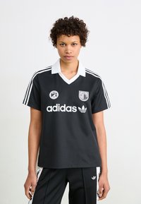 Melnā futbolista krekla ar baltajiem akcentiem, V veida apkakle, trīs svītras uz piedurknēm un logo uz krūtīm. Audums izskatās gluds un elpojošs.