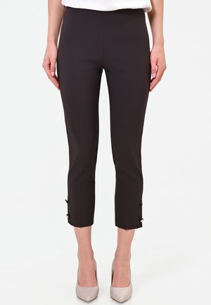 Donna che indossa pantaloni neri corti con dettagli a bottoni vicino alle caviglie e tacchi alti chiari su uno sfondo semplice.