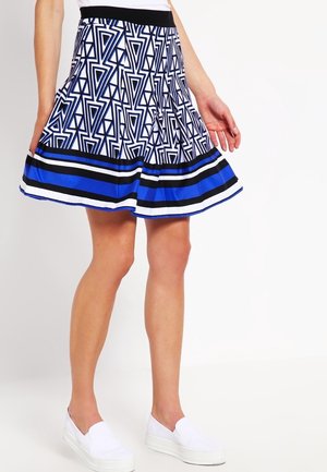 Vrouw in een geplooide rok met zwart, wit en blauwe geometrische patronen en witte instappers, staande tegen een effen witte achtergrond.