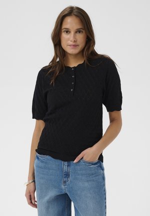 Nő fekete rövid ujjú kötött felsőt visel, gombokkal és kék farmert, egy kézzel a zsebében egy egyszerű háttér előtt áll.