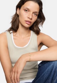 Ljusbeige linne med vit kant, parat med mörkblå jeans. Modellen bär ett guldarmband med hänglåsdetalj.