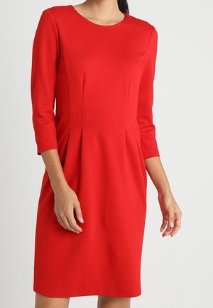 Vrouw die een knie-lange, nauwsluitende rode jurk draagt met driekwart mouwen en een ronde halslijn, staande tegen een effen achtergrond.
