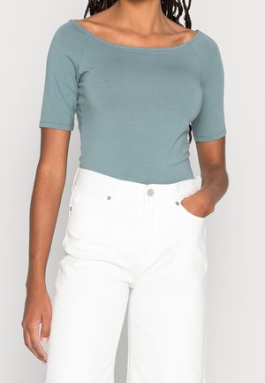 Vrouw draagt een teal off-shoulder top met korte mouwen en witte hoge taille broek, met één hand in de zak tegen een effen achtergrond.