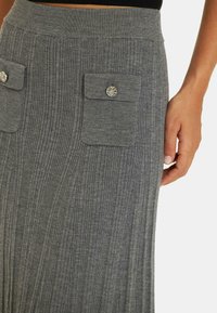 Jupe en maille côtelée grise avec une taille ajustée, présente deux poches avant avec des boutons décoratifs en argent, texture lisse et lignes verticales.