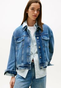 Giubbotto di jeans in azzurro chiaro, con chiusura a bottoni, tasche sul petto e una vestibilità rilassata, indossato sopra una camicia a righe azzurra chiara.