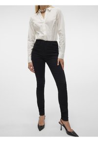 Femme portant une chemise blanche à manches longues boutonnée, un pantalon skinny noir, des escarpins noirs pointus et un grand collier de perles argenté.