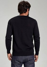 Pull en maille noir à manches longues et poignets côtelés, avec un col rond classique et une coupe ajustée au niveau de la taille.
