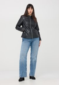 Giacca in pelle nera con colletto rialzato, chiusura zip, tasche laterali e dettagli delle cuciture, abbinata a jeans azzurri svasati.