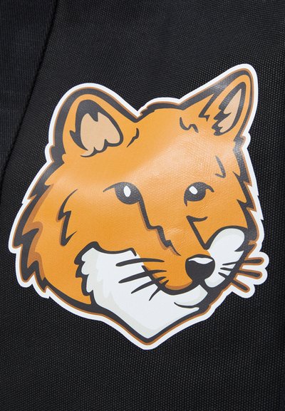 Autocollant en forme de tête de renard orange avec des contours noirs et des accents blancs, appliqué sur un fond en tissu noir texturé.