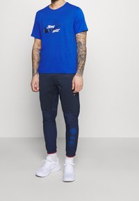 Blaues Baumwoll-T-Shirt mit bedrucktem Logo, kombiniert mit dunkelblauen Jogginghosen mit strukturierten Akzenten und Stickereien. Weiße Sportschuhe.