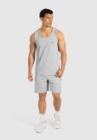 Uomo in canotta grigia senza maniche e pantaloni corti abbinati, in piedi su uno sfondo neutro, con calze bianche e sneakers.
