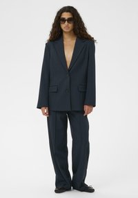 Marineblauer, maßgeschneiderter Anzug aus glattem Stoff; verfügt über einen einreihigen Blazer mit breiten Revers und locker sitzende, gerade geschnittene Hosen.