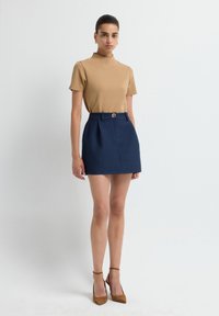Top corto a maniche corte color beige abbinato a una mini gonna blu scuro a motivo, con una vita dotata di un bottone rotondo dorato e pieghe. Scarpe beige con tacco alto.