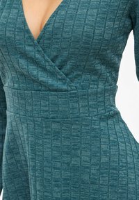 Teal knit dress featuring a V-neck and long sleeves. The fabric has a textured pattern with vertical lines and a fitted waist.