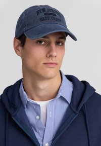 Cappello blu navy con ricamo "NEW JERSEY EAST COAST", in tessuto di cotone, con visiera curva, indossato con un hoodie blu navy e una maglietta a righe sotto.