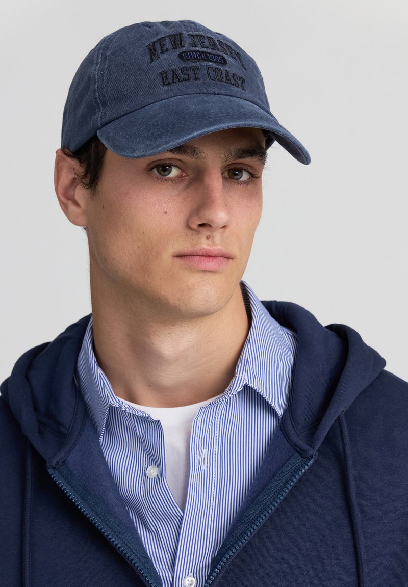 Cappello blu navy con ricamo "NEW JERSEY EAST COAST", in tessuto di cotone, con visiera curva, indossato con un hoodie blu navy e una maglietta a righe sotto.