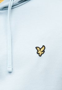 Ανοιχτό μπλε φούτερ με κουκούλα, με μικρό κίτρινο κέντημα πουλιού με μαύρο περίγραμμα στο στήθος, διαθέτει κορδόνι κοντά στη λαιμόκοψη.