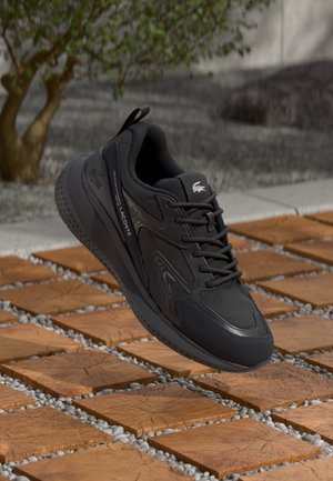 Baskets noires Lacoste avec logo sur la languette et le côté, posées au-dessus d'un chemin en carreaux de bois avec de petits cailloux entre les carreaux et un arrière-plan flou.