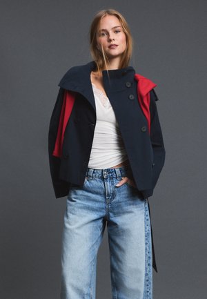 Jeune femme portant une veste bleu marine et rouge par-dessus un haut blanc bordé de dentelle et un jean bleu clair ample, debout devant un fond gris.